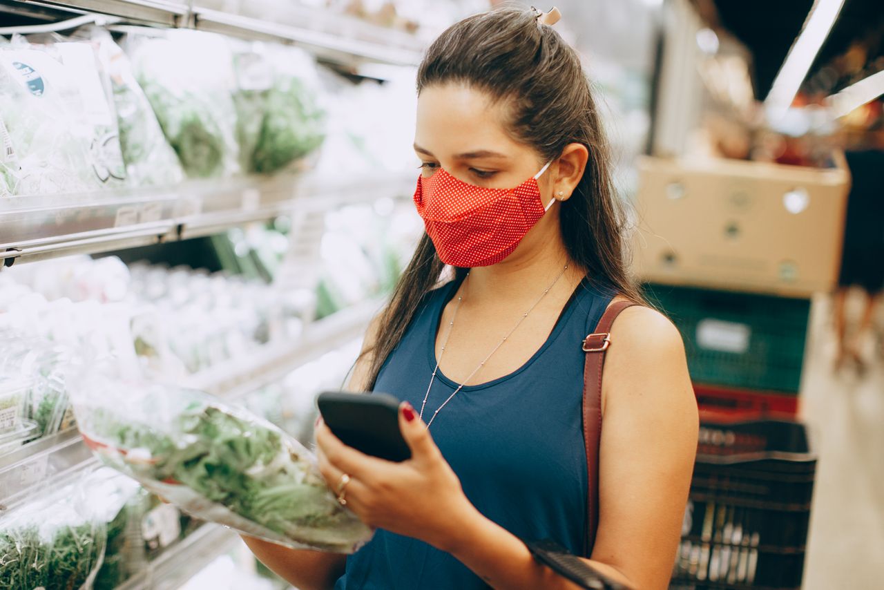 services-05 Woman wearing red face mask shopping for greens in a supermarket, checking her phone.