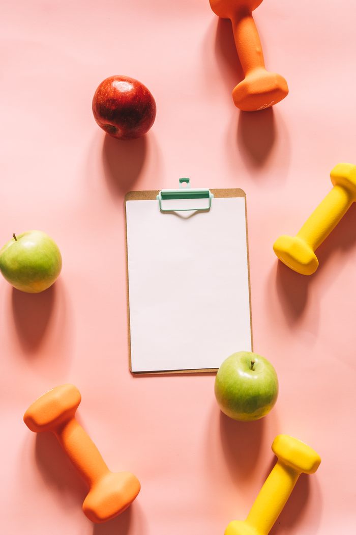 mission-02 Top view of colorful apples, dumbbells, and clipboard on a pink background, ideal for health and fitness themes.